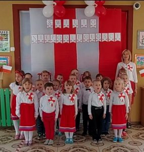 Grupa dzieci uroczyście ubranych wraz z nauczycielką stojących na tle materiałów biało czerwonych z napisem Święto Niepodległości
