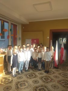 Na obrazku stoi grupa przedszkolna dzieci ubranych odświętnie. Obok dzieci widoczne flagi Polski.