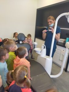 Pani stomatolog trzyma w ręku sztuczną szczękę i szczoteczkę do zębów, prezentuje dzieciom jak należy prawidłowo myć zęby.