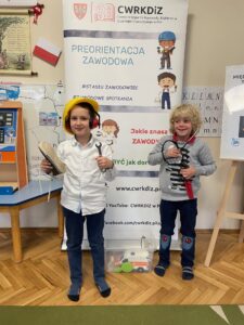 dwóch chłopców stoi przed plakatem trzymają w rękach atrybuty różnych zawodów